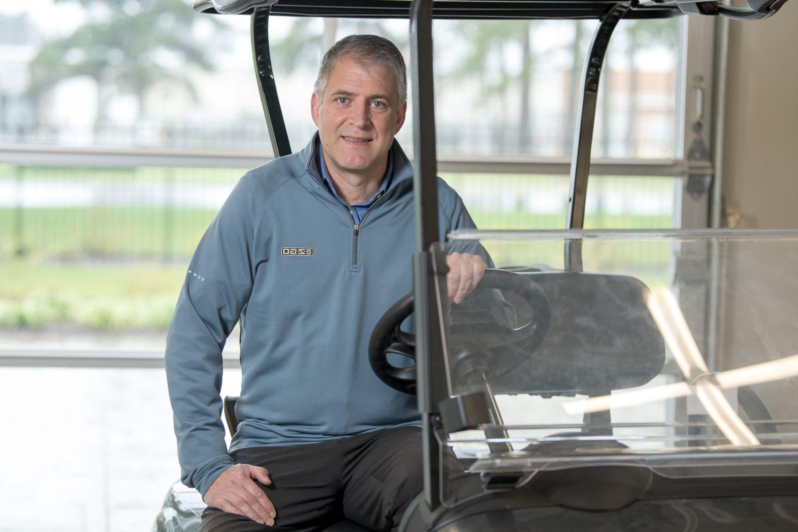 Mr. Gunnar Kleveland, a Caucasian male, sits at the wheel of a golf cart wearing a blue fleece pullover and grey slacks while smiling at the camera.