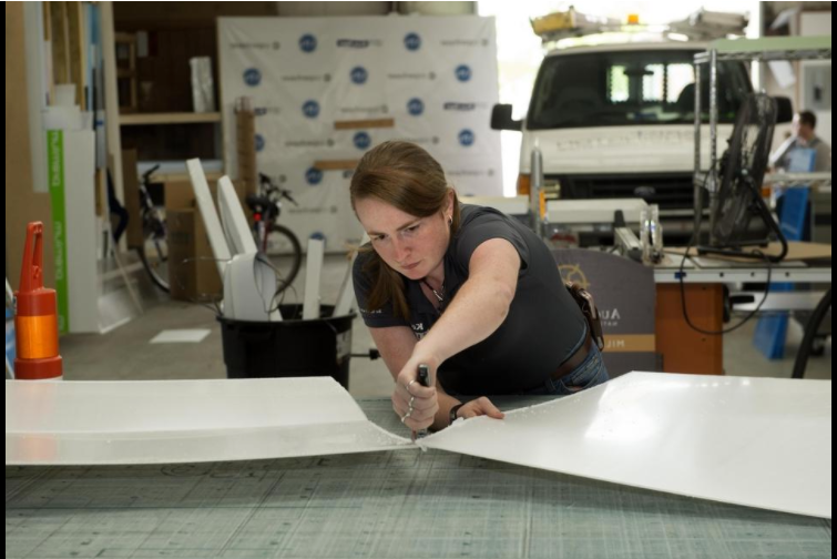 A Caucasian female with brown hair, Brittany Doster is leaning over a table while cutting a white board. A white van with design supplies and tools are in the background.