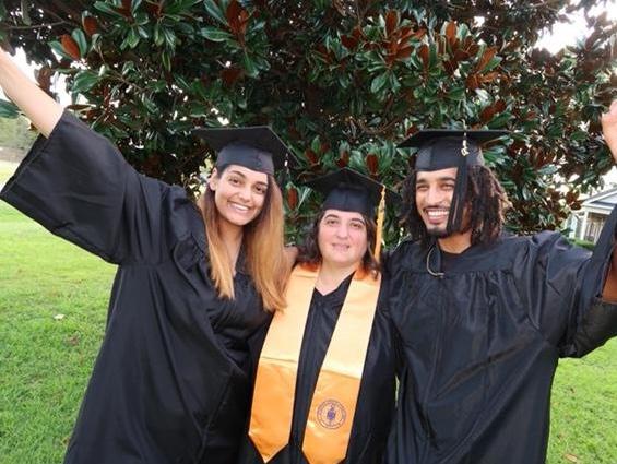 Smiling African American male with dreadlocks wearing a black graduation gown, black graduation cap with gold tassel, gold necklace and a blacks shirt; hand is spread out; center: Caucasian female with brown hair wearing a black graduation gown, black graduation cap with a gold tassel and a gold stole with a logo, left: smiling, female with golden brown hair wearing a black graduation cap, black graduation gown; hand is spread out; group is standing in front of a tree