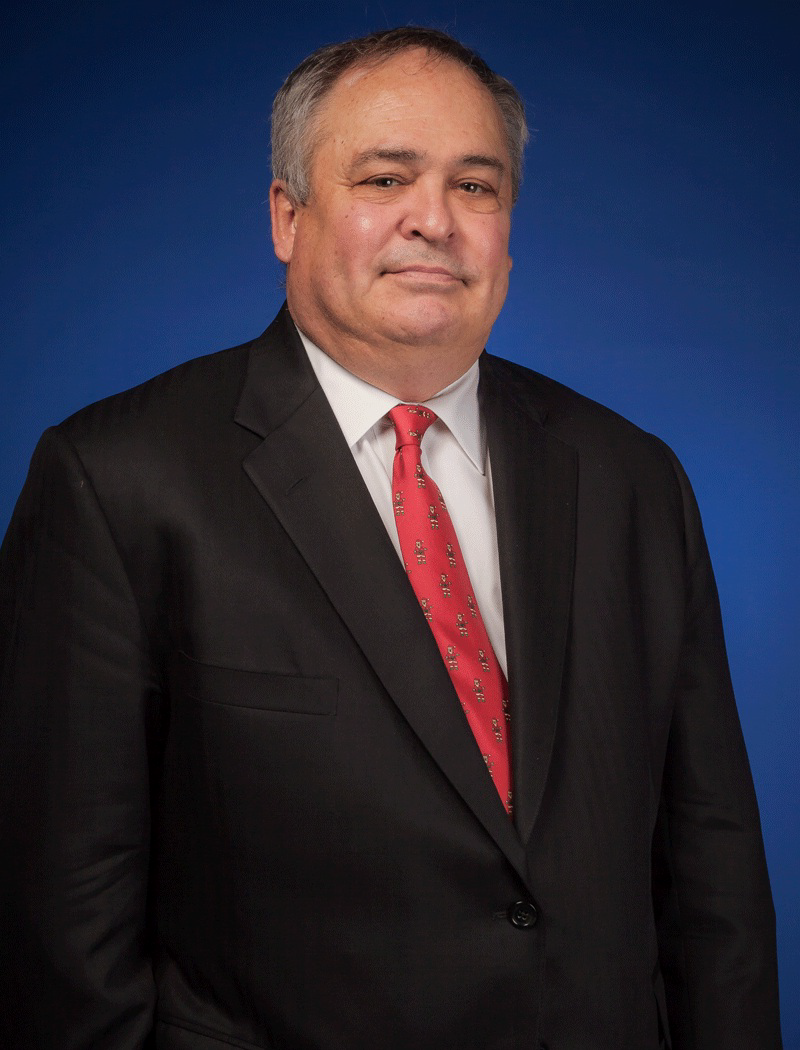Smiling Caucasian male wearing a black suit, red tie, white collared shirt, posing for a photo against a dark dark blue background