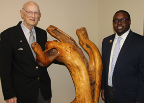African American male, wearing black glasses, a blue suit, white collared shirt, light blue tie, silver Augusta Tech lapel pin, standing next to a wood statue; Caucasian male wearing glasses, a black suit, blue collared shirt, pocket square; both are posing for a photo against a tan-colored wall