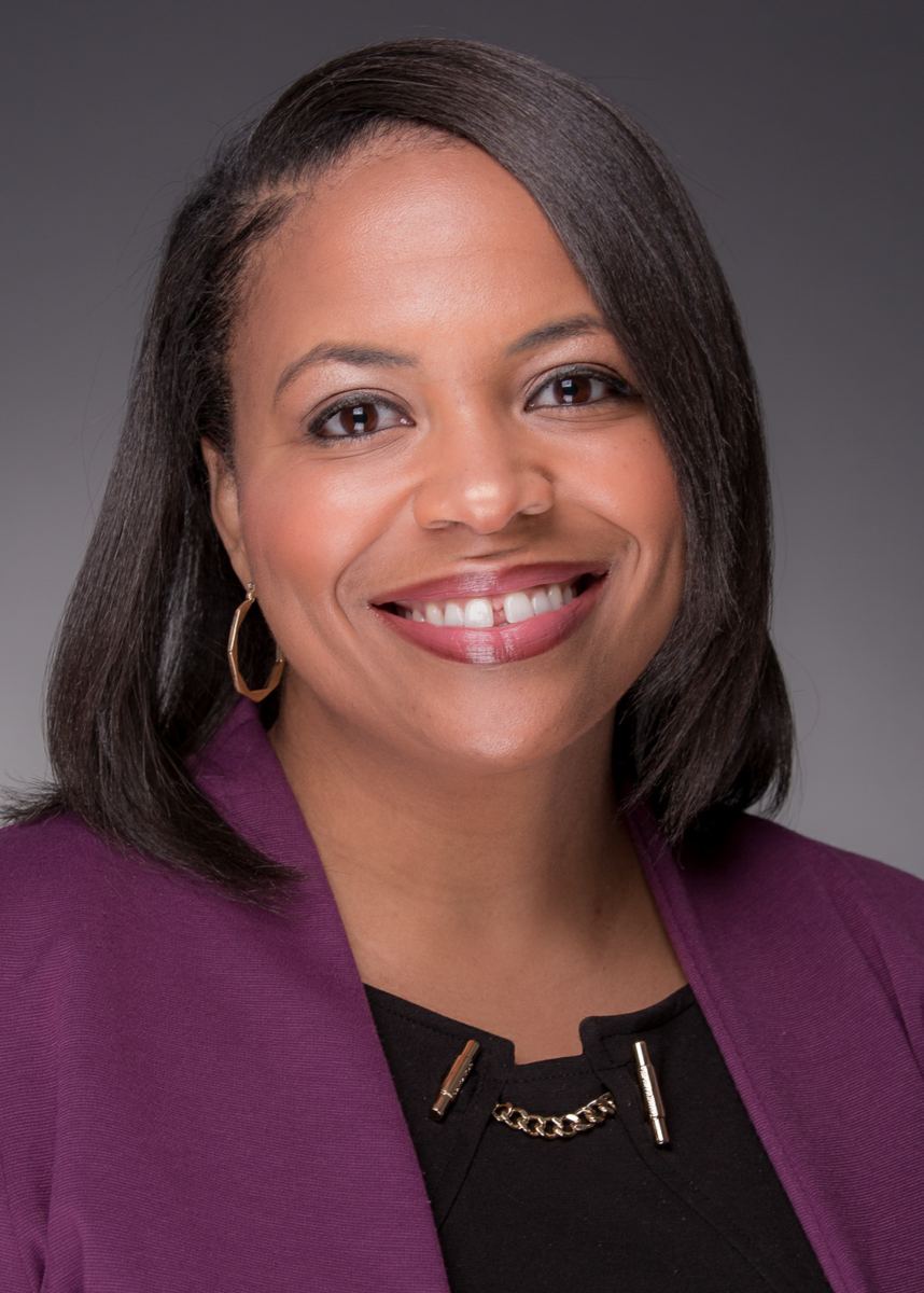 Ms. Christine Ball, an African American female with shoulder length dark brown hair, smiles at the camera wearing a purple suit jacket and a black dress shirt with a bronze metal clasp on the front and gold hoop earrings.