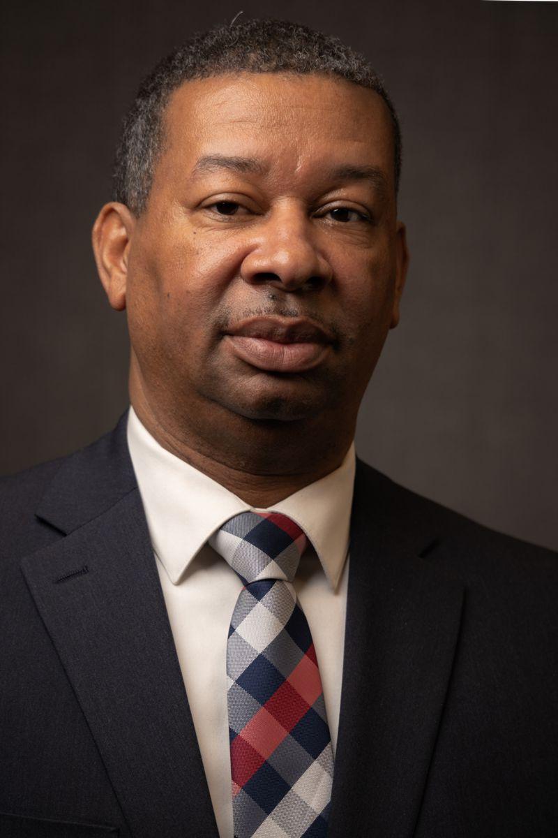 Dr. Howie Gunby, an African American male wearing a grey suit with a white collared shirt and a grey and white plaid tie with a single stripe of red.