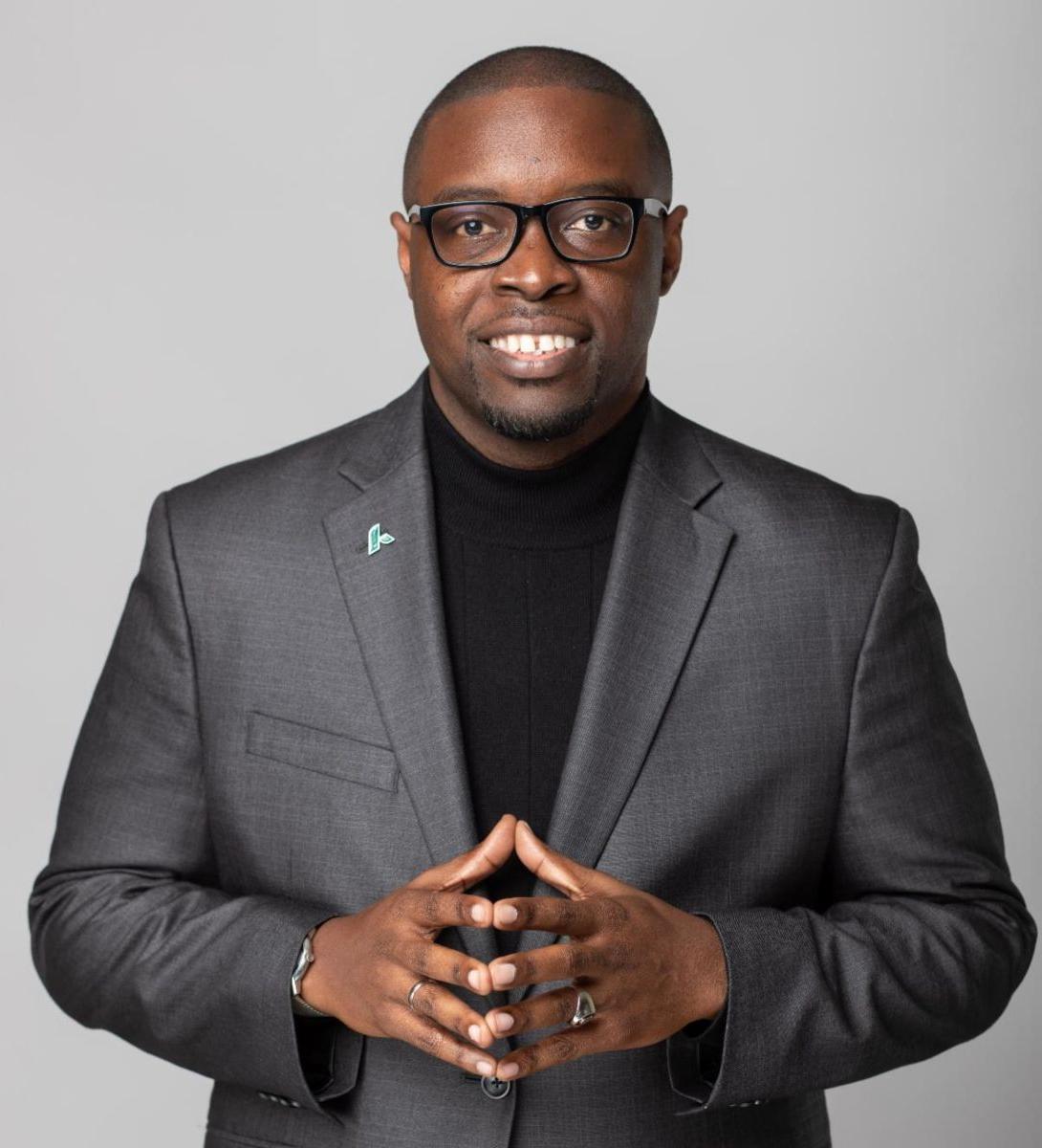 Dr. Jermaine Whirl, an African American male, smiles wearing a grey suit jacket over a black shirt with his hands steepled towards the camera in front of his abdomen.