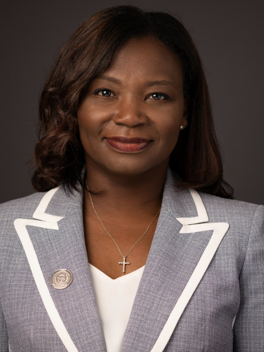 Tara Jenkins, an African American female with shoulder length wavy brown hair, smiles wearing a grey suit jacket with white bordering, a white dress shirt and a gold cross necklace.