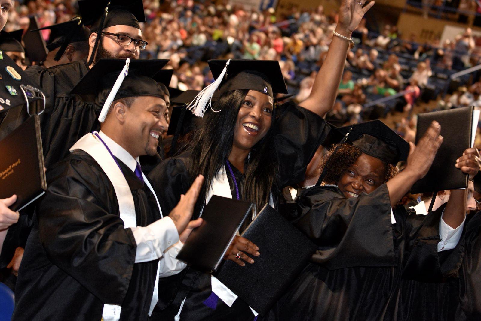 森林的绿色, 超链接, 白色的技术信用证书按钮, 粗体, 在文字下面加了下划线，上面是一群拥挤的毕业生和家人，他们的注意力集中在四名戴着黑色毕业帽、穿着白色流苏长袍和披肩的毕业生身上:一名非洲裔美国女性微笑着，左手举着一个黑人文凭/学位托, 在她的左边，一位非洲裔美国女性微笑着举起戴着金属手镯的右手，左手拿着她的黑人学位证书, 一位非裔美国男性微笑着站在那里，右手拿着他的黑人文凭/学位证书，左手举在后面, 一个留着黑胡子戴着眼镜的白人男性站在他们三人的右边.