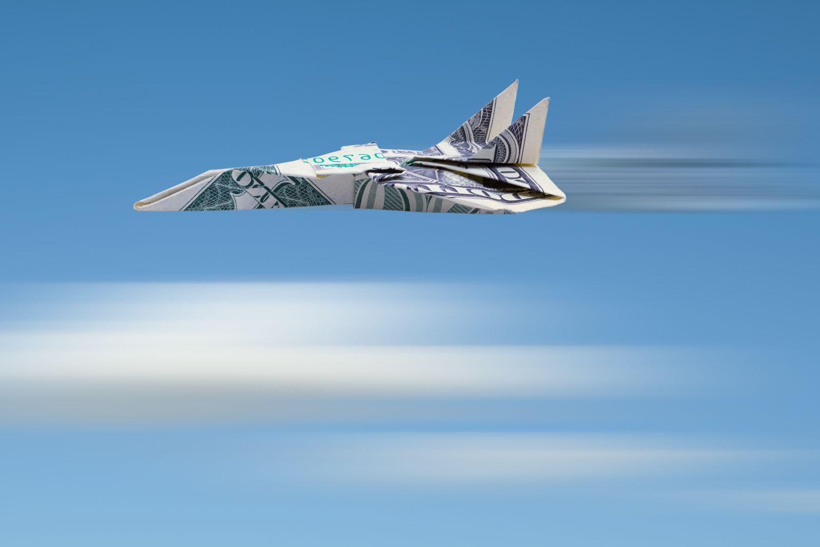 用1美元纸币制作的纸飞机以天蓝色为背景，下面的白色模糊线看起来像模糊的云，引擎后面的黑白模糊线代表飞机在高速飞行.