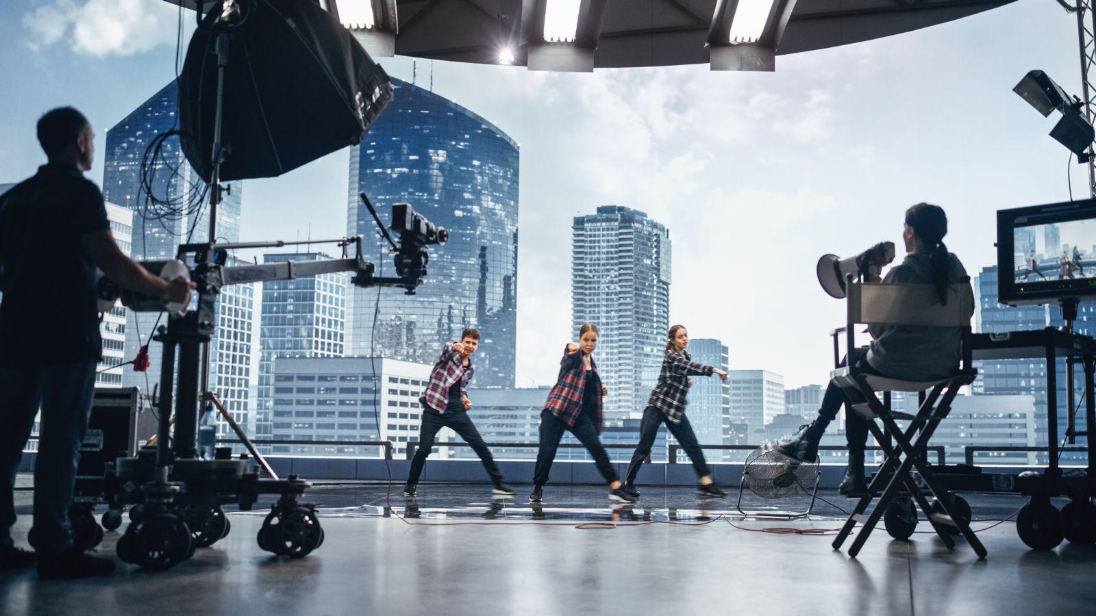 Three dancers are being filmed against a city backdrop in the background with a director, film crew, and equipment in the foreground.